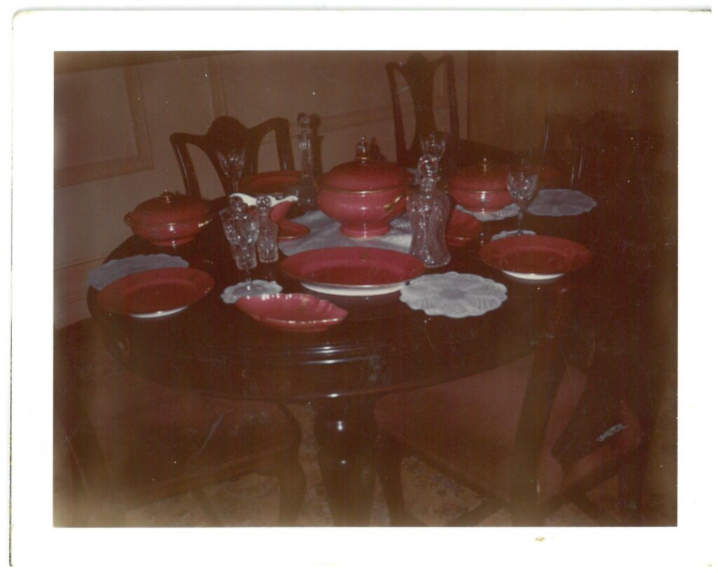 A slightly faded photograph showing a selection of pink dinner service pieces. It's dark pink - with gold on the rims and handles. Faintly visible (not clear details) is a family crest