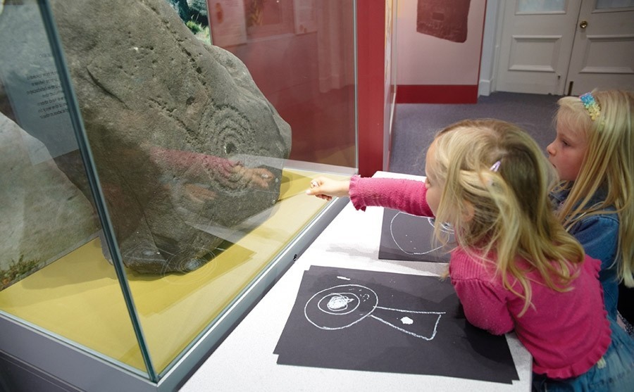 2 children pointing at an old fossil behind a glass case.