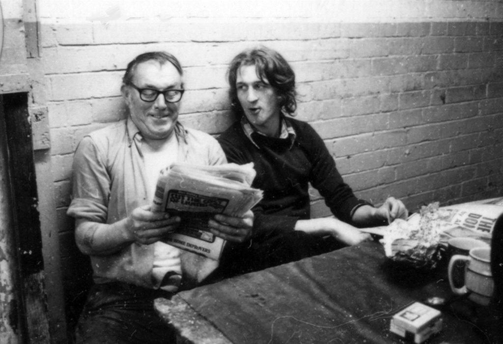 Ian sitting in the work canteen, next to am older man. Ian is in dark black jumper with long hair, the other worker is in a white shirt and light coloured cardigan, holding a newspaper. Table in view in foreground