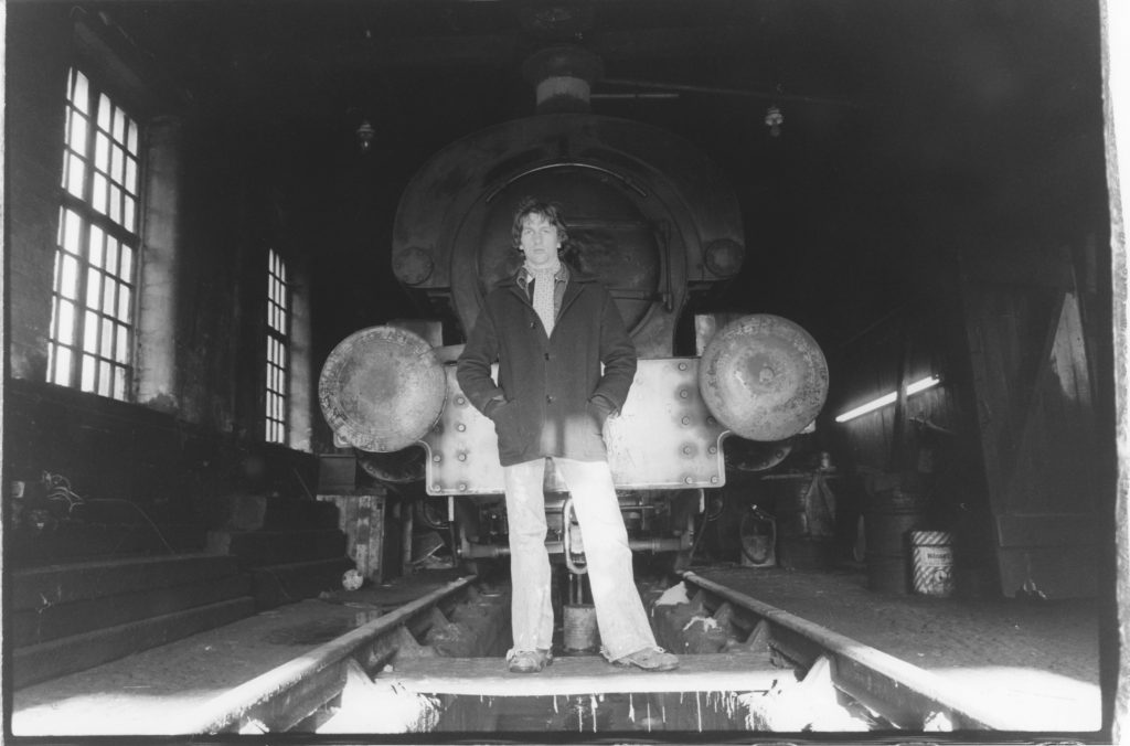 black and white photograph. Ian is posed (still with shoulder length hair) in front of a train, standing on a board straddling the rail cutting. It's inside a big train shed