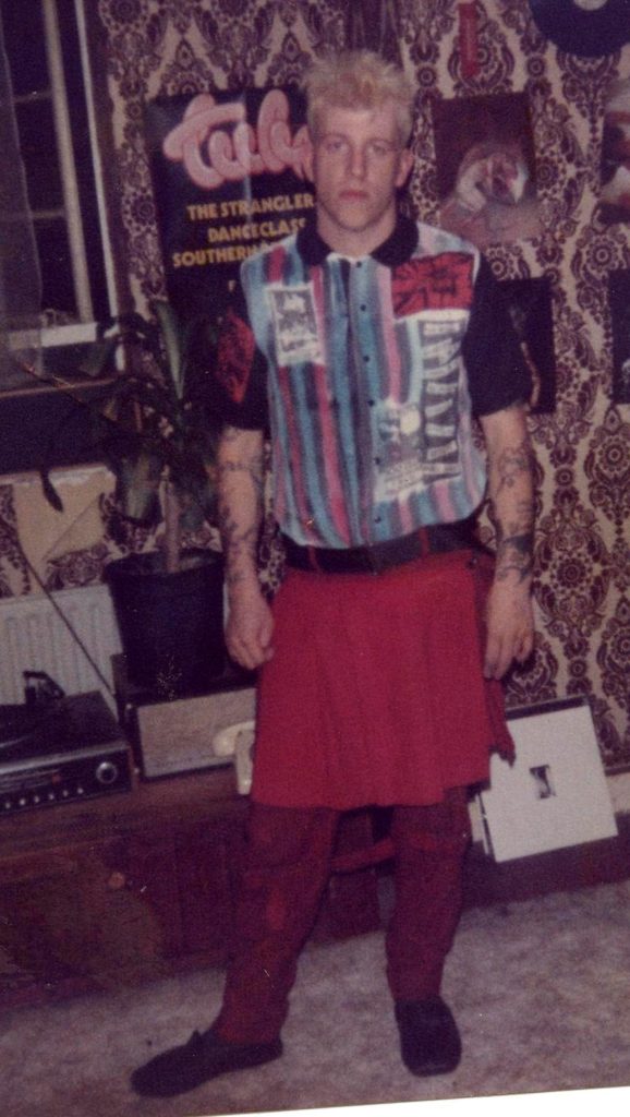 Young man, with bleached blond hair, dressed in red trousers, a red skirt over, and a stripey 'punk' shirt, with various slogans. Stood in front of a record player on chest, Record LP's visible to the right. Various posters visible behind - including 'Tuba' - with the Stranglers visible at top of list