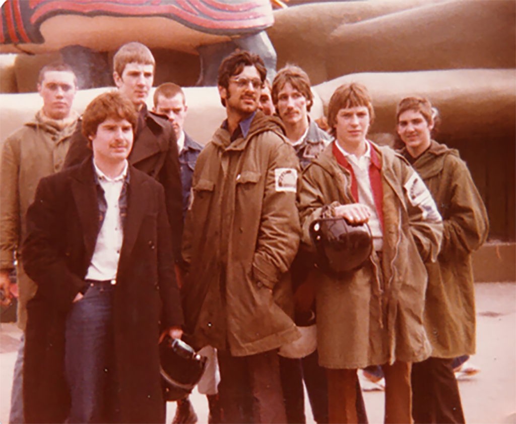 Image of a group of young men - 'Scooterboys'- the majority are young white men, but the central figure is a young asian man. They are all dressed in typical 'scooter boy' fashion.