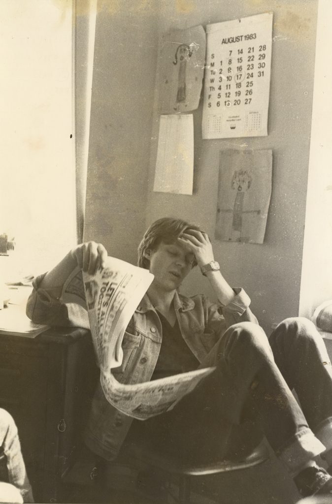 B&W image of a young man, dark jeans, light coloured jacket sat in chair reading. Various images pinned to wall behind him including an calendar dated August 1983