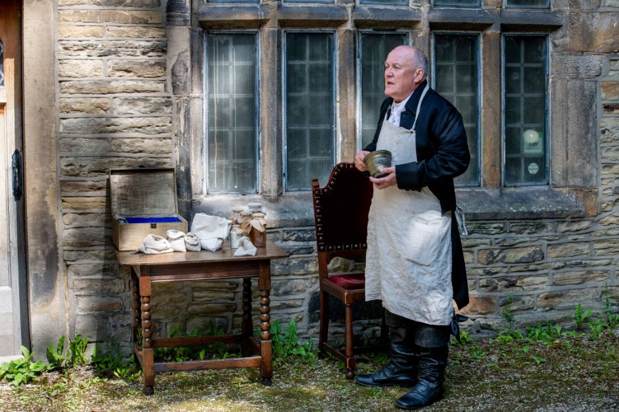 actor dressed as an apothecary, holding a mortar and pestle,  a table with medicines is to the side