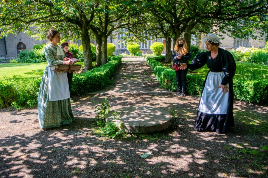 2 women dressed in old fashioned Victorian looking clothes in the middle of a woodland area.