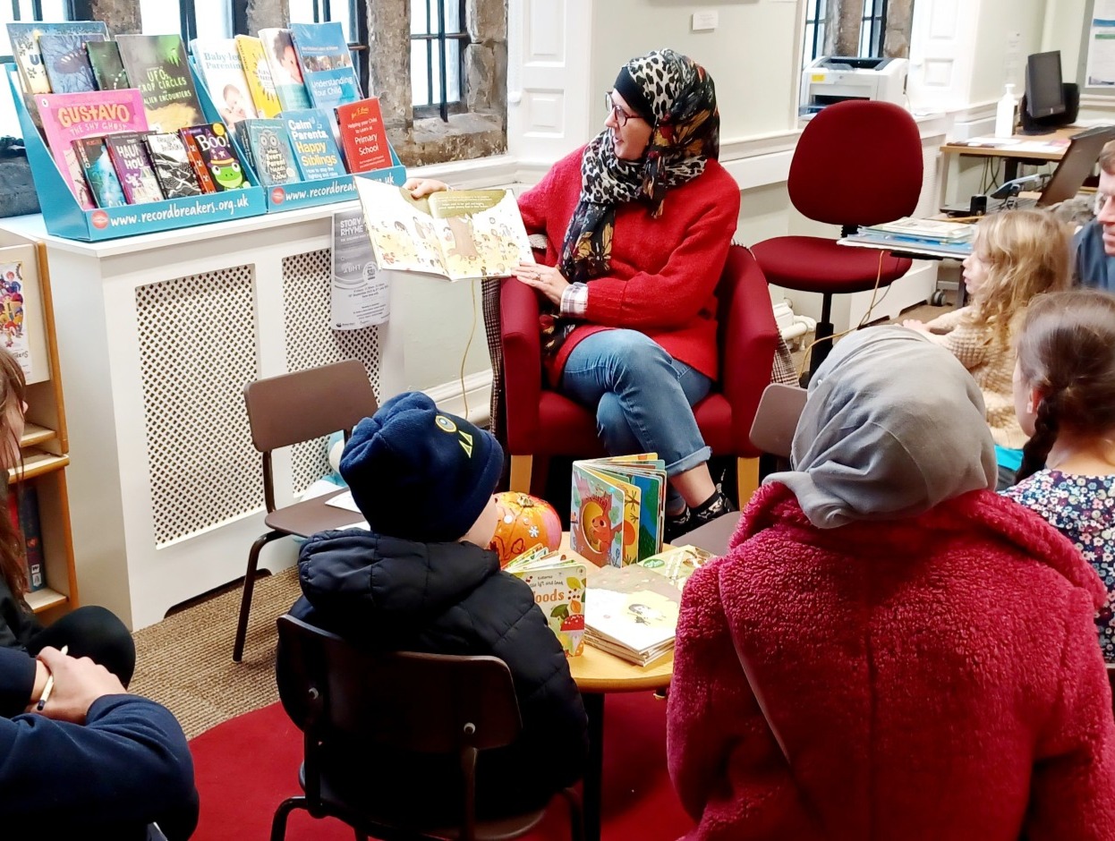 a volunteer sat in a chair in the library, reading a story to a group of families
