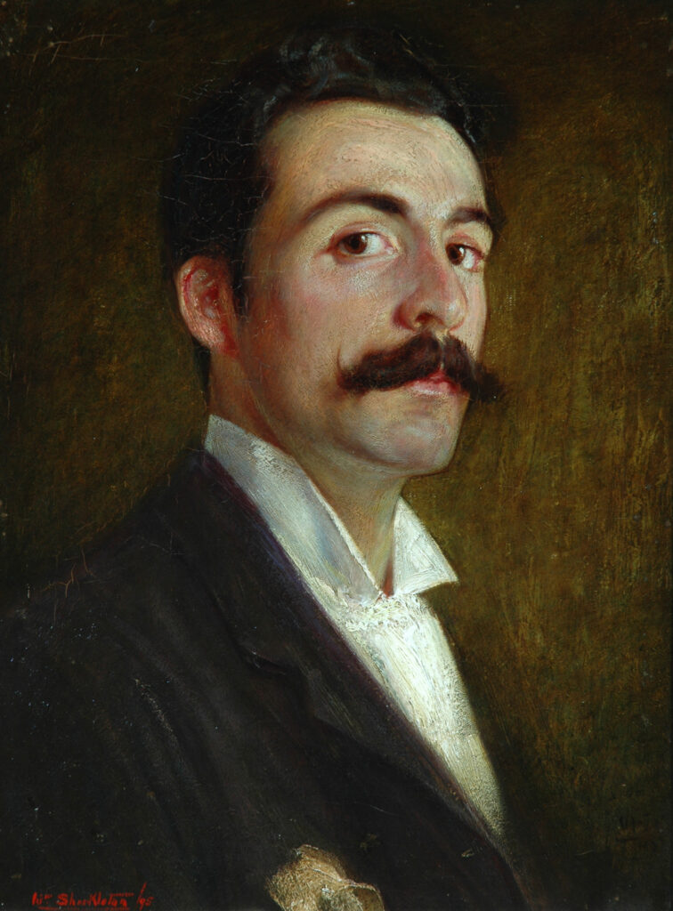 Image of a youngish man (mid twenties?)  Dark brown hair, and a rather splendid mustache.  He has dark brown eyes, and is wearing a black suit with white shirt