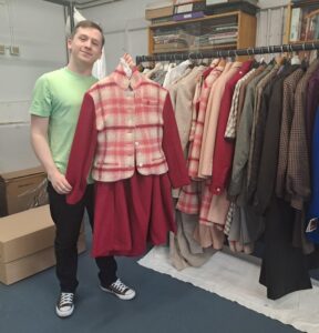 Callum stood next to the Woolmark collection on a clothes rail, holding up a Wendy Dagworthy suit comprising jacket and skirt in pink and cream tones.