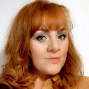 A red headed woman with long wavy hair wearing a sandy coloured top