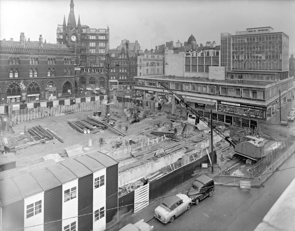 Photograph shows the construction of Arndale House, Bradford, on the former site of the Swan Arcade. There is a construction site which the foundations of a new buildings being built. The Wool Exchange building is visible behind it. Photographer: C H Wood, 1963. In Bradford District Museums and Galleries collection (57533).