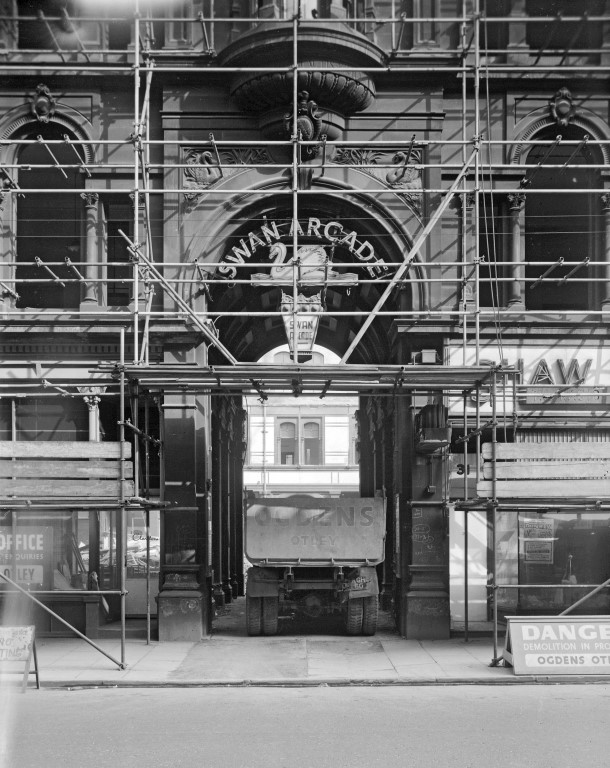 Photograph shows the Swan Arcade, Bradford, just before its demolition. It was an ornate Victorian building – the photograph shows its arched entrance to the arcade with a sign saying ‘Swan Arcade’ with scaffolding all on it and a construction truck driving through the arch into it. Photographer: C H Wood, 1962. In Bradford District Museums and Galleries collection (140818).