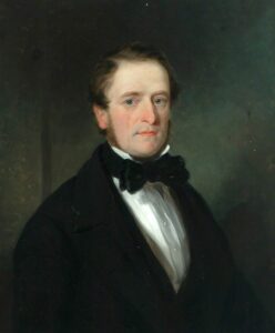 Cleanshaven white man with dark brown hair and side burns. He has blue eyes, and is wearing a black suit, white shirt and black bowtie. He is looking out towards the viewer