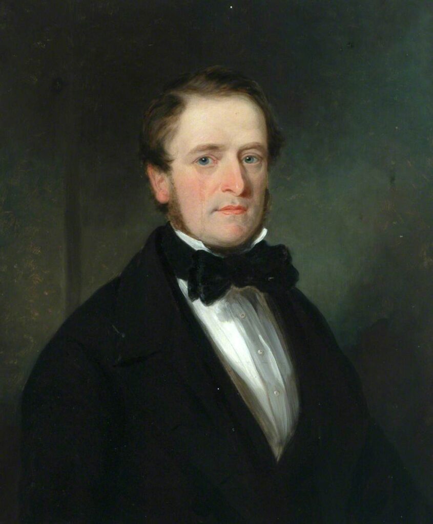 Cleanshaven white man with dark brown hair and side burns.  He has blue eyes, and is wearing a black suit, white shirt and black bowtie.
He is looking out towards the viewer