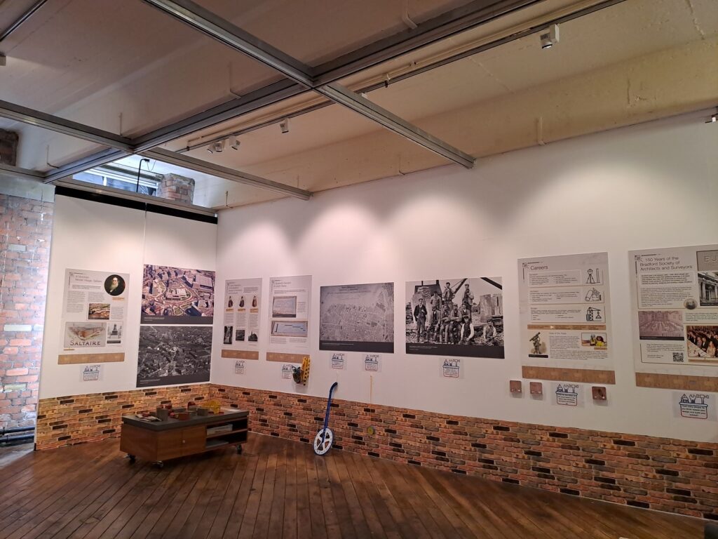 Photograph shows one side of the exhibition with panels on white walls and a brick-effect wallpaper border underneath. There is toy town set with a train track and block buildings for younger visitors set out for them to be town planner.