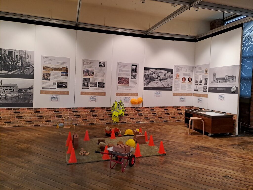 Photograph shows the other side of the exhibition with panels on white walls and a brick-effect wallpaper border underneath. There is area of children’s toy set out with foam bricks, cones, a wheelbarrow and hard hats like a construction zone.