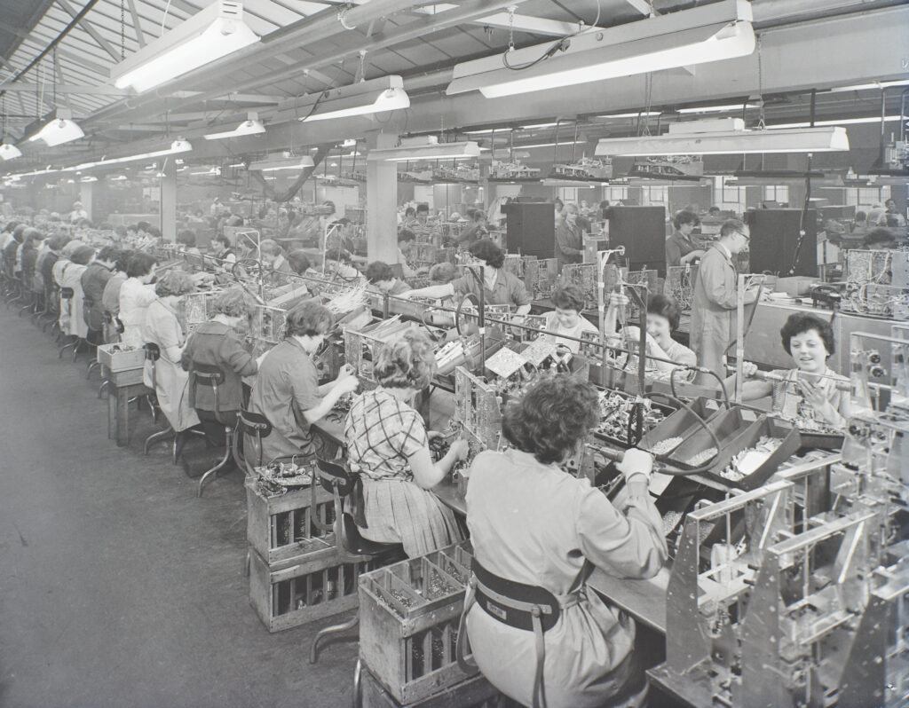 B&W photo of women working at an assempbly line in a larger building.  They are wearing a range of outfits, rather than a uniform.  They have lots of different components sitting in boxes in front of them and most the women are holding what looks like soldering irons