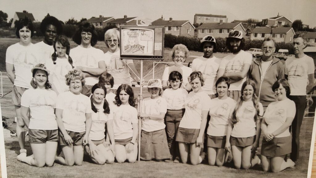 Group employees in shorts and t-shirts and very 70s haircuts, stood around a pole with a cardboard cut of a televison, ready to compete