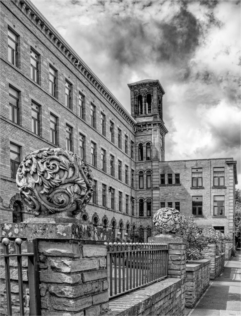 Legrams Mills.  Shows the mill building behind, with stone wall and decorative iron railings in front - the pillars for the wall are topped with round stone balls carved with twining plants