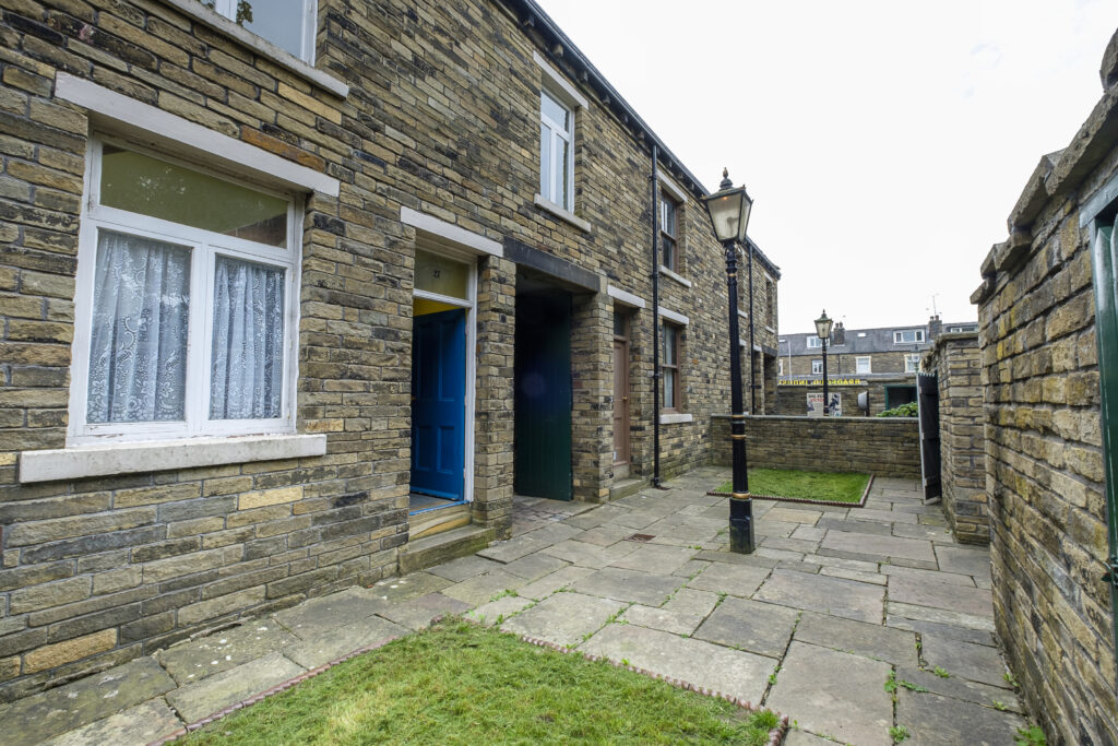 the exterior of a row of back to back houses.  Shows a ginnel (through alley) under one of the windows, and a lampost in the foreground.