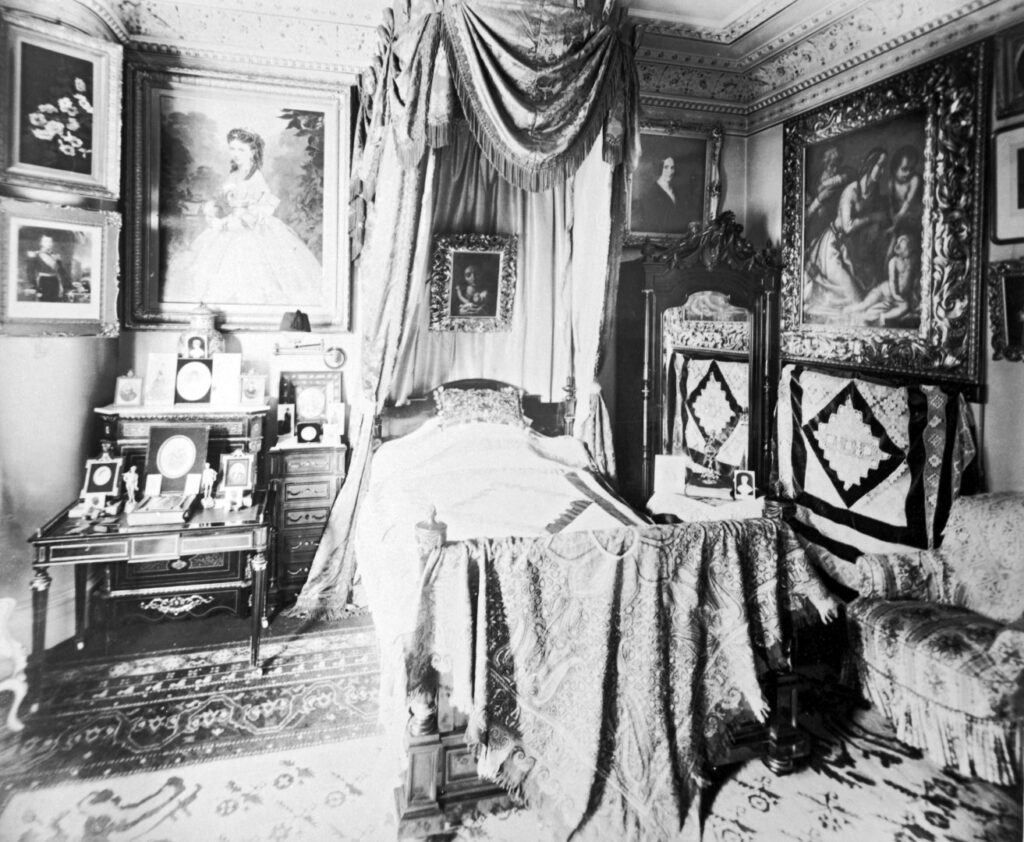 B&W photo showing a bedroom.  to the left is a portrait of Marie Louise Butterfield, underneath that desk covered in various photos, minatures and painting.  In the centre is a bed with curtained canopy.  To the right is additional furniture, paintings and an armchair