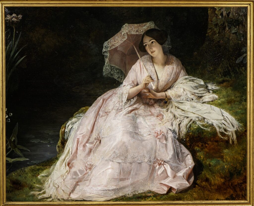 A lady with dark hair, and a long, 1830s/40s style dress in light pink.  She is sat reclined on the bank of stream, which is just visible in the bottom left corner, and she is holding a matching pink parasol