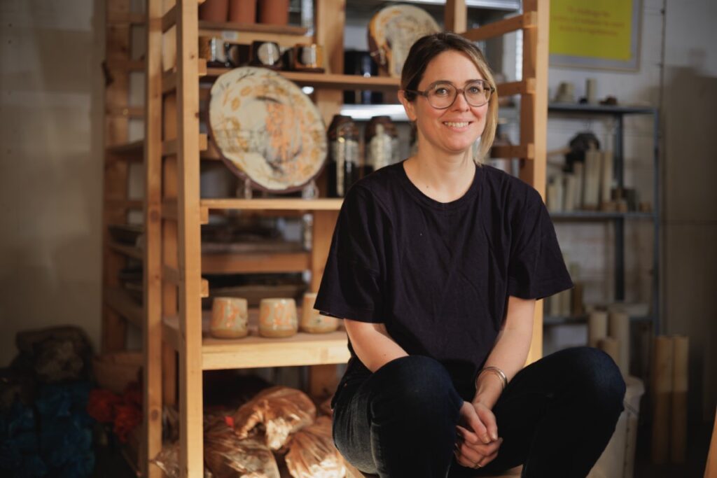 A blond haired woman wearing glasses, and dressed all in black sat in front of a rack of ceramics,  with more visible in the background