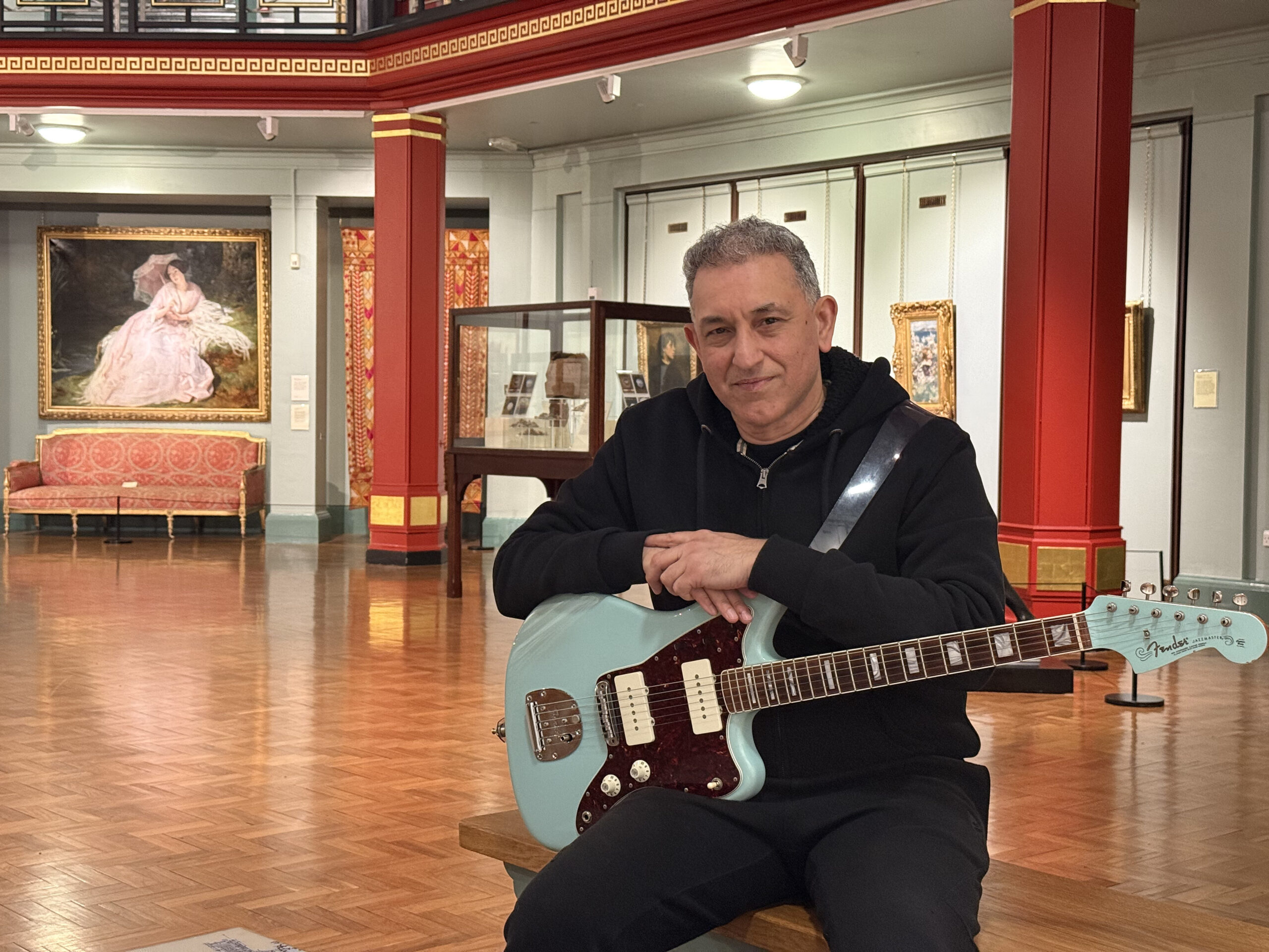 Aziz Ibrahim sat in the Bracewell Smith Hall, with his guitar on his lap and a painting visible in the background