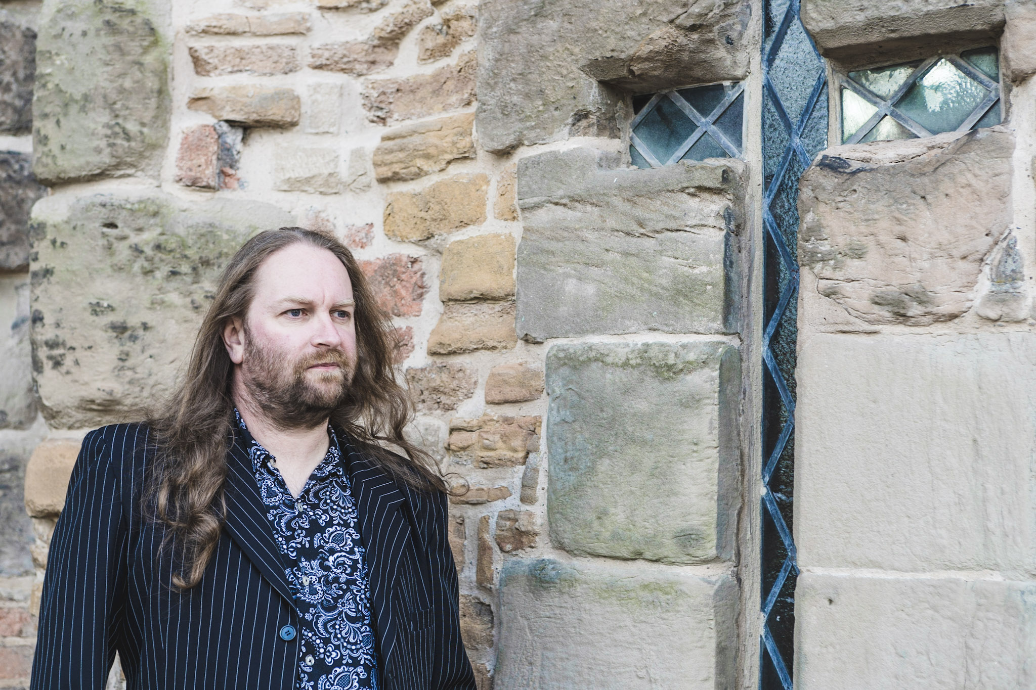 Dr James Wright, a white mane with long reddish brown hair standing pensively by an architectural feature on a historical building