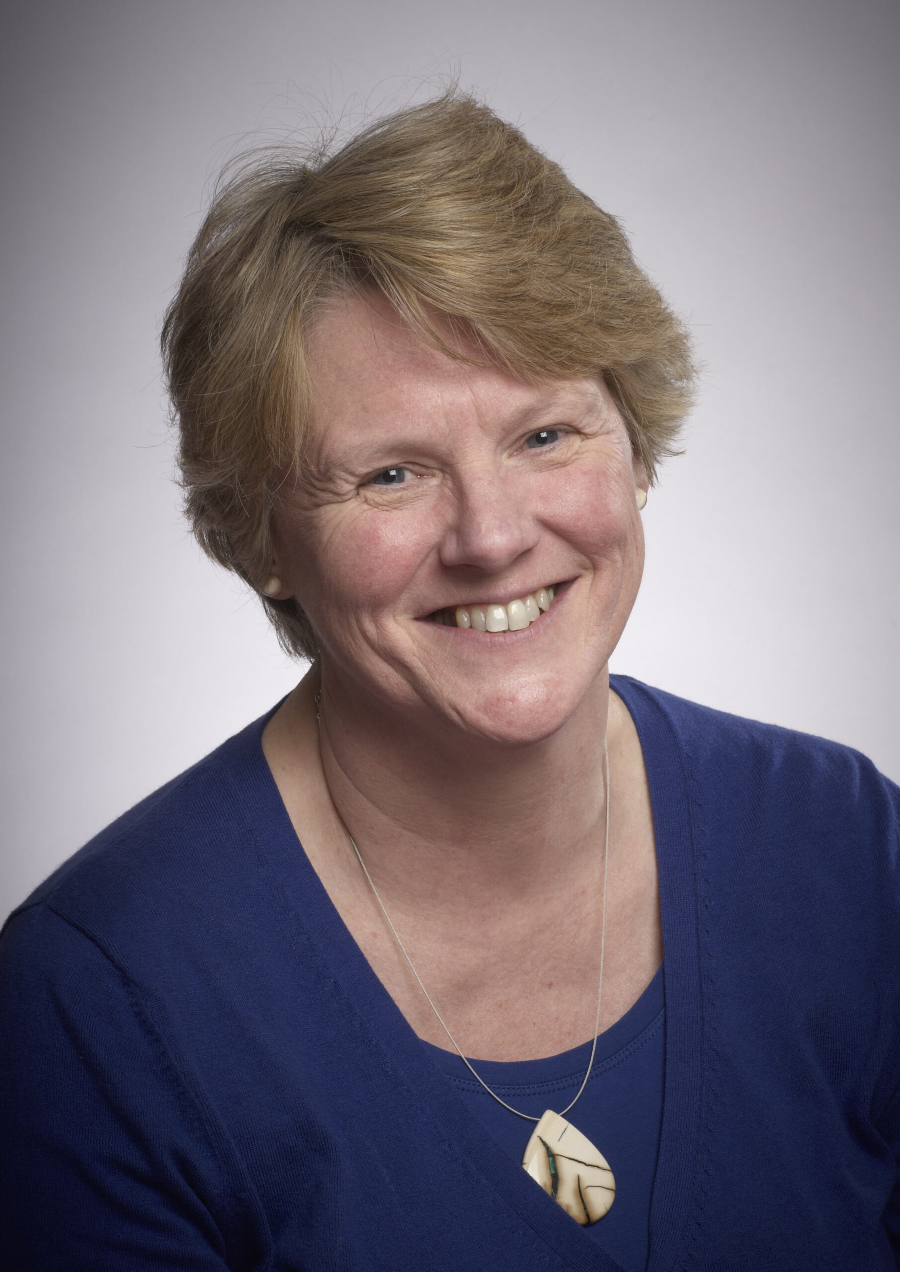 image of Jill Cook. A white woman with short blonde hair, and is wearing a blue top and a carved pendant necklace