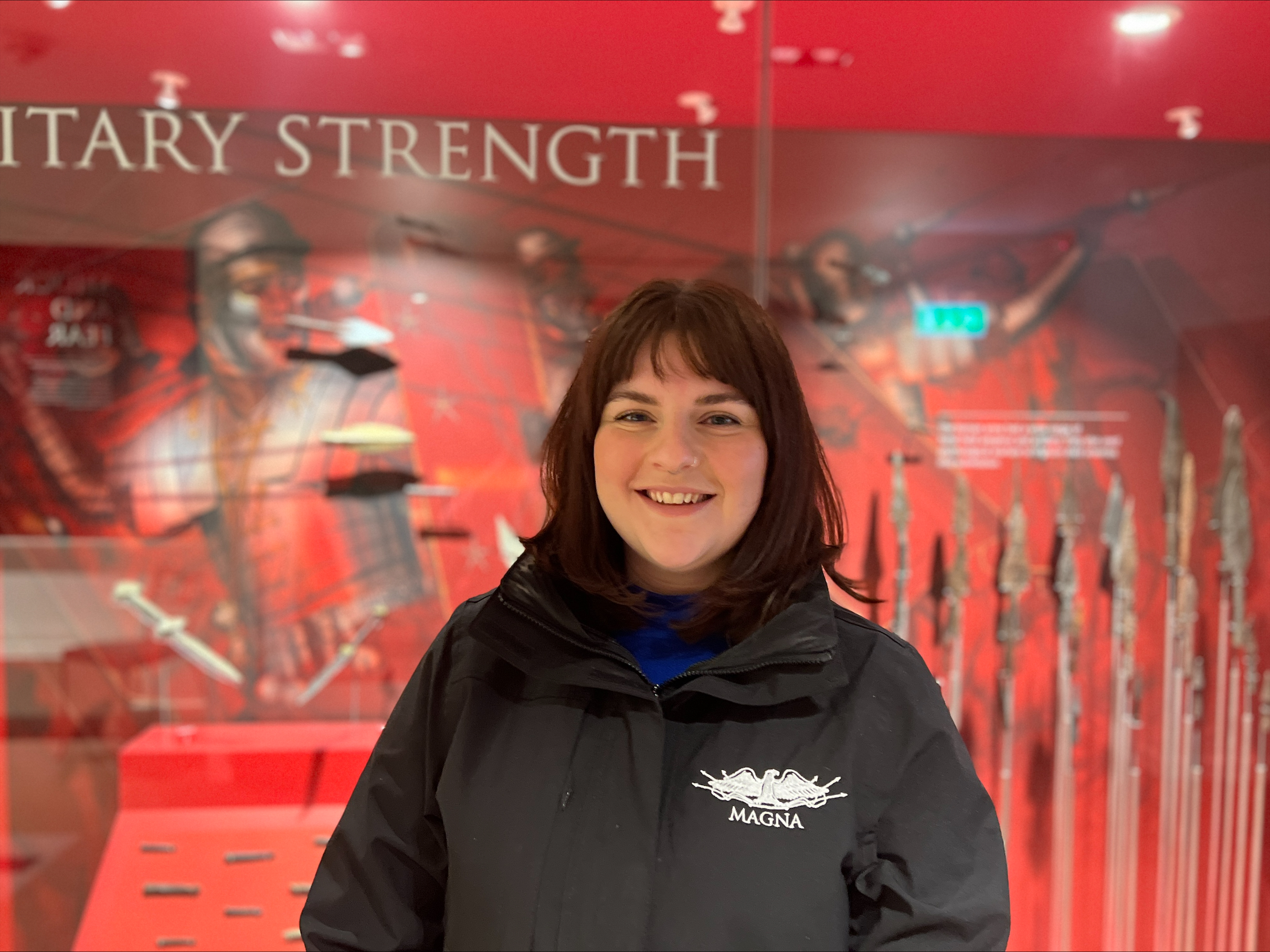 a dark haired woman standing in front of a case of archaeological objects, with 'Vindolanda' embroidered on her jacker
