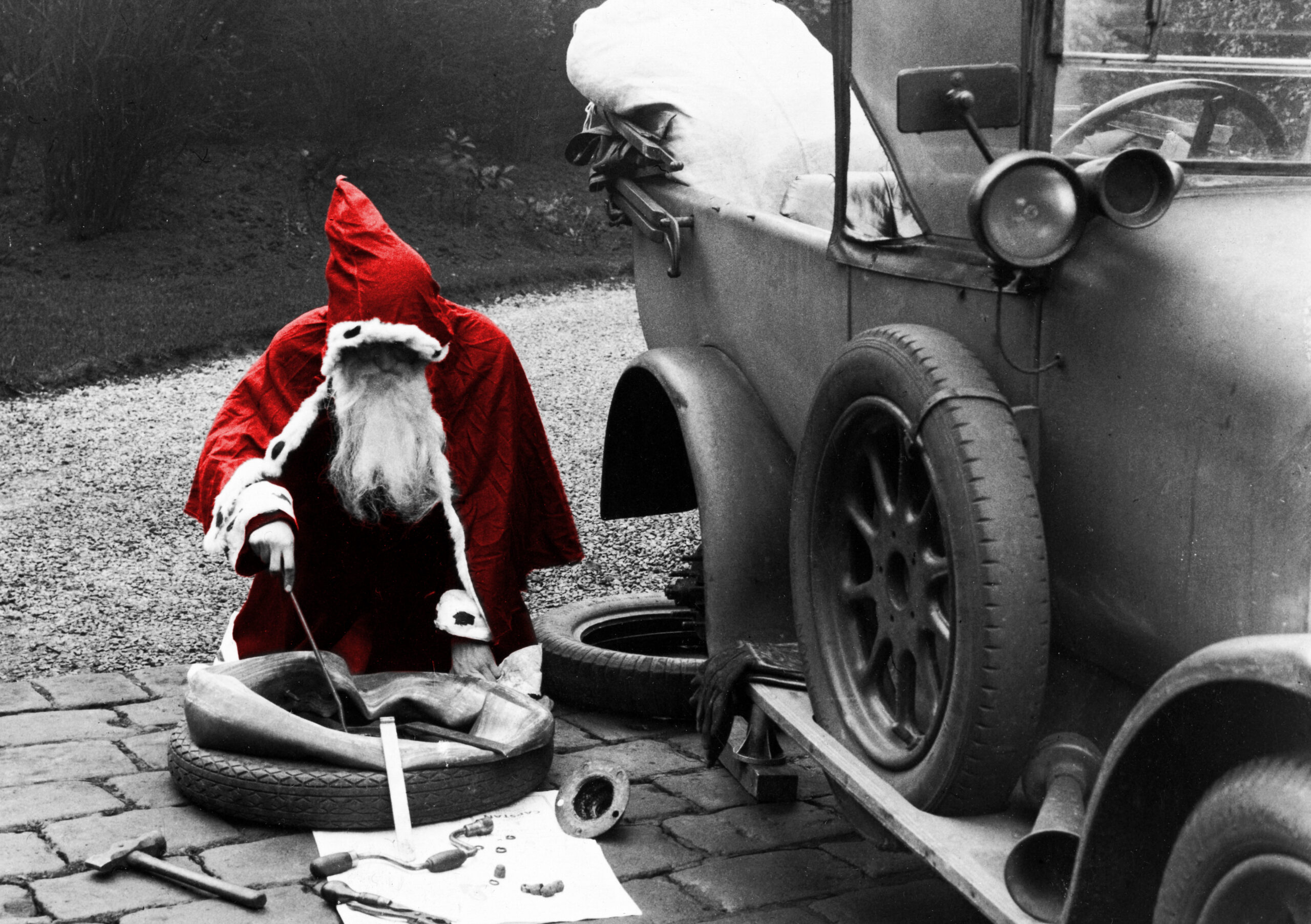 Mostly B&W image- father christmas is colourised in red. He is in the middle of changing a tire on an old car