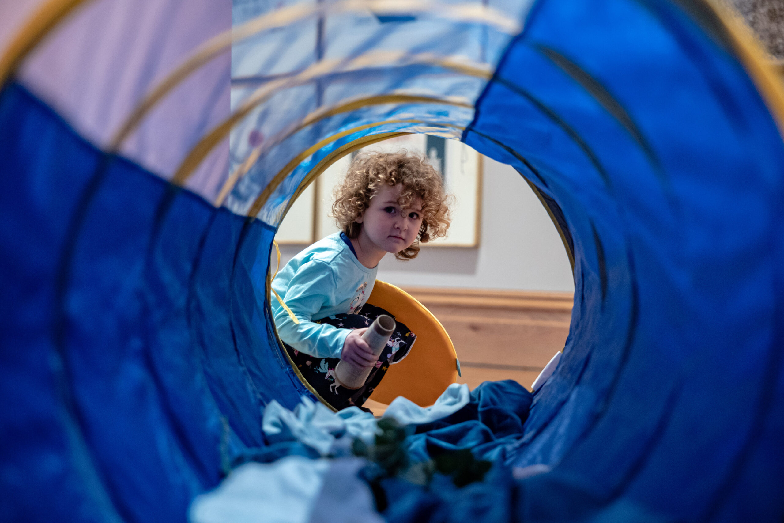 Small curly-hair child peering through a fabric tunnel, artwork in the background