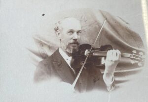 B&W photo of a man with a violin posing in front of draperies (head and shoulders only) He has balding hair and a full beard and mustach