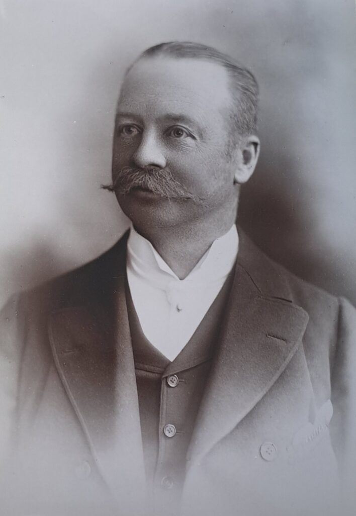 a white man in victorian clothing - dark suit, coat. White tie and collar.  He has darkish hair, swept back, slightly receding.  No beard, but a fine moustache...