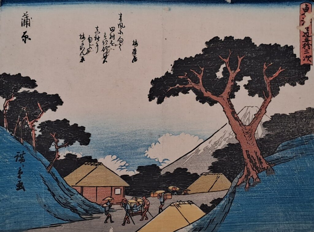 a wood block print Japanese in style.  shows a village with people, with trees and houses in the foreground and a mountain behind