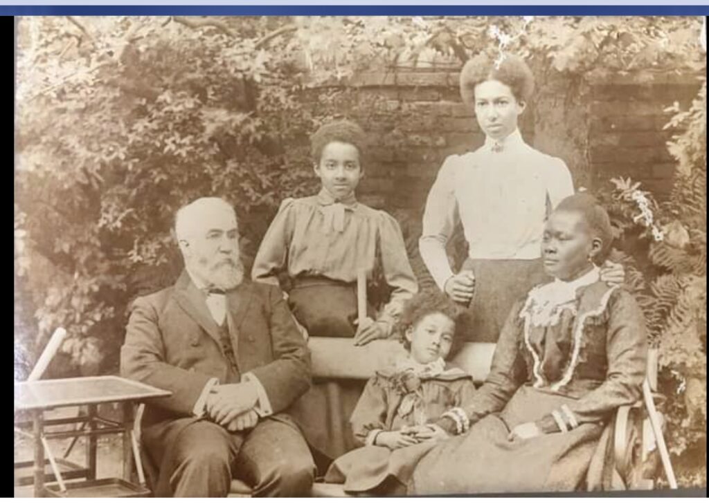 family photograph - father (a white man with white hair and beard) is seated on a bench onthe left, the mother (a black woman with dark hair) is seated on the right of the bench. The two oldest children (daughters) are stood behind the bench and the youngest (a boy) is sat between his two parents