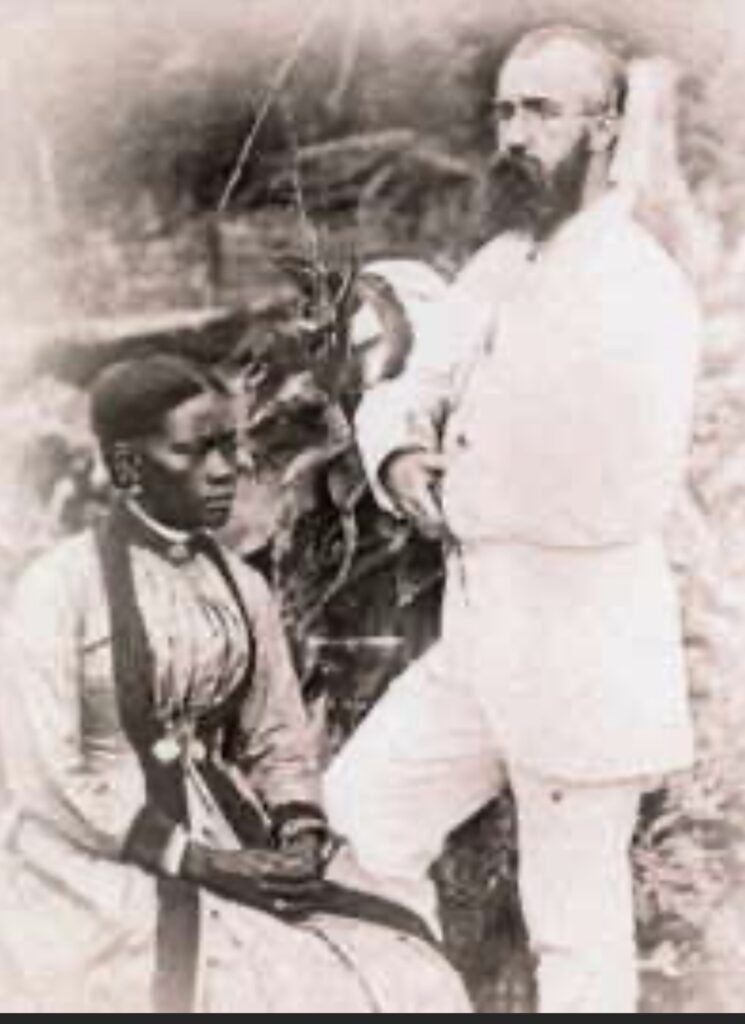 image of a black woman sat down - light coloured dress with darker trim, and a white man standing next to her. He has a light colured suit on, and a large beard