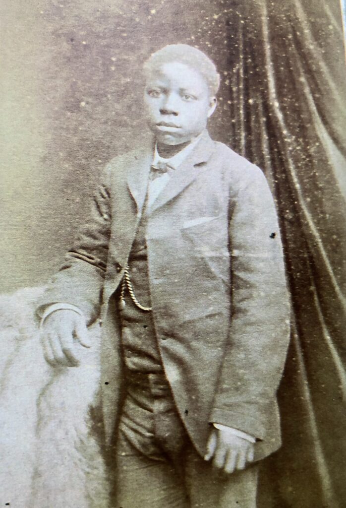 a young black man (early teens perhaps?) he is dressed in a formal suit -dark coloured, white shirt. standing in a formal studio setting