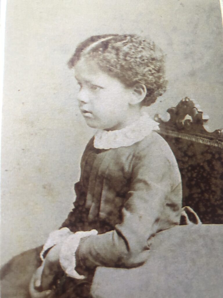 B&W photo of a young black child in formal victorian dress. Dark coloured dress, light coloured dress collar (possibly lace?) sat formally on a chair in a studio set up