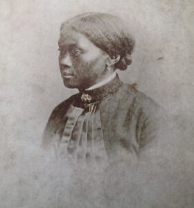 B&W photo. A head and shoulders photo of a young black woman. Her hair is pulled back, she is wearing a dark coloured (black?) dress with a high collar and brooch. She is in profile rather than face on.