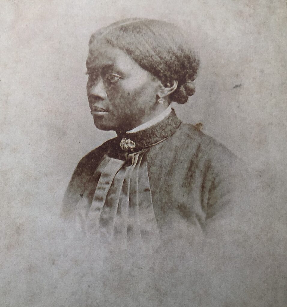 B&W photo. A head and shoulders photo of a young black woman. Her hair is pulled back, she is wearing a dark coloured (black?) dress with a high collar and brooch. She is in profile rather than face on.