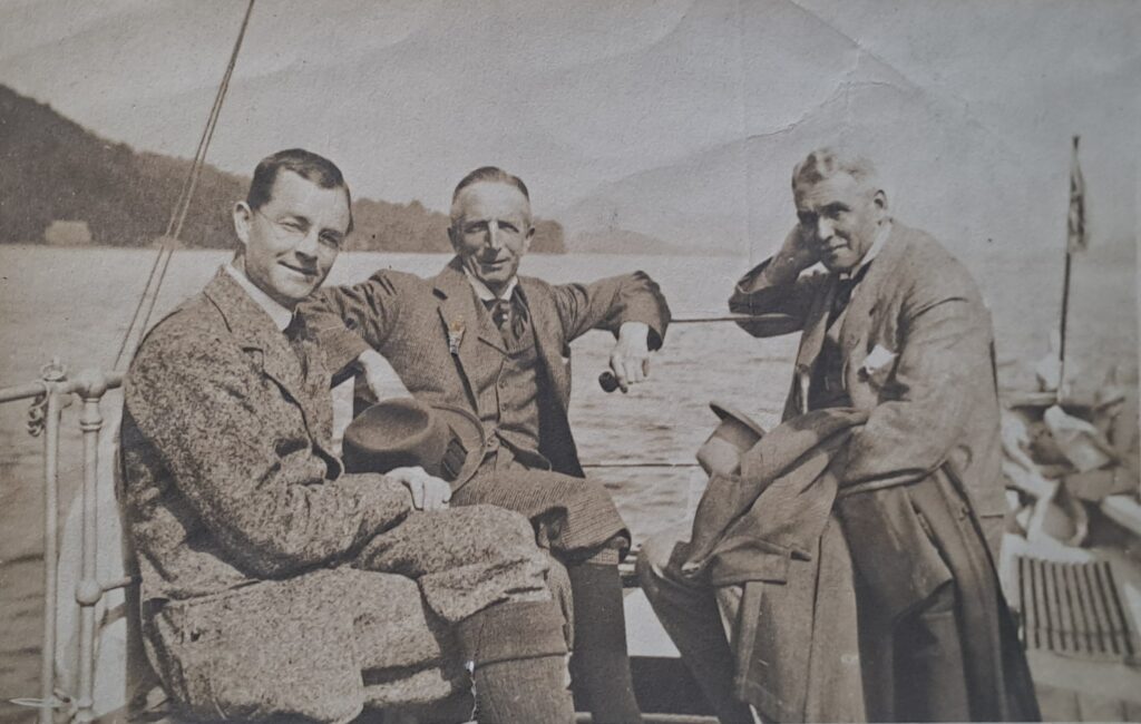 a group of three white men on a boat on a lake.  They're all wearing 'country' tweed type suits, sock etc suitable for countryside activities