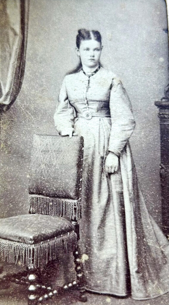 a white woman, in a light coulured dress, Victorian in style. She is standing behind a chair in a photographers studio set. It is a full length image