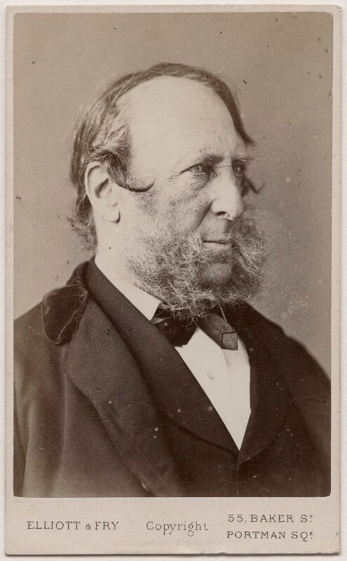 A sepia-toned Victorian-era portrait photograph showing a person wearing a dark formal jacket, a white shirt, and a bow tie. The image includes a visible beard and sideburns. At the bottom of the photograph, the text reads ‘ELLIOTT & FRY Copyright 55, Baker St Portman Sq.