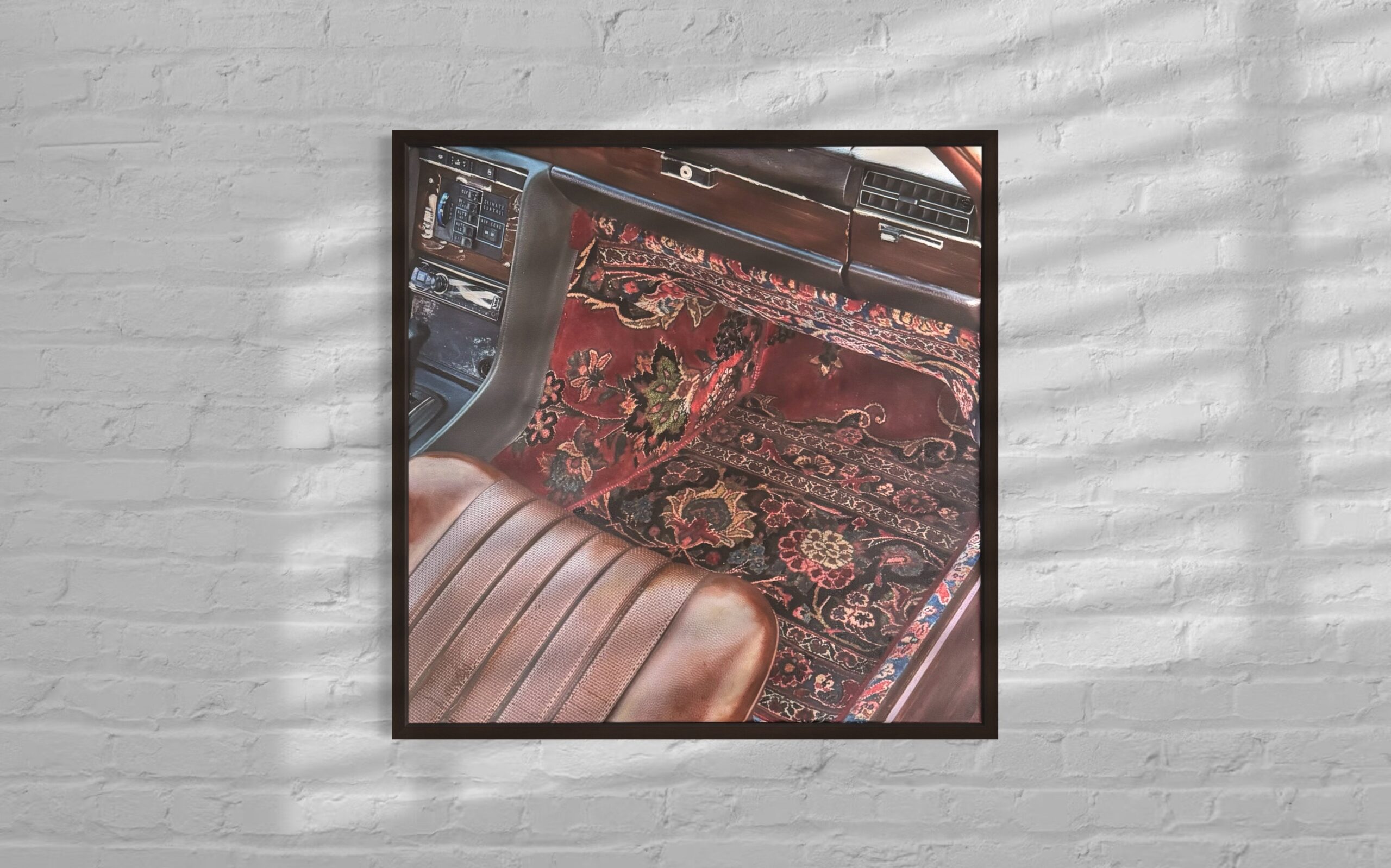 Framed artwork depicting the interior of a vintage car with a brown leather seat and dashboard, featuring an ornate red and black Persian-style rug laid across the floor. The frame is mounted on a white brick wall with soft shadows