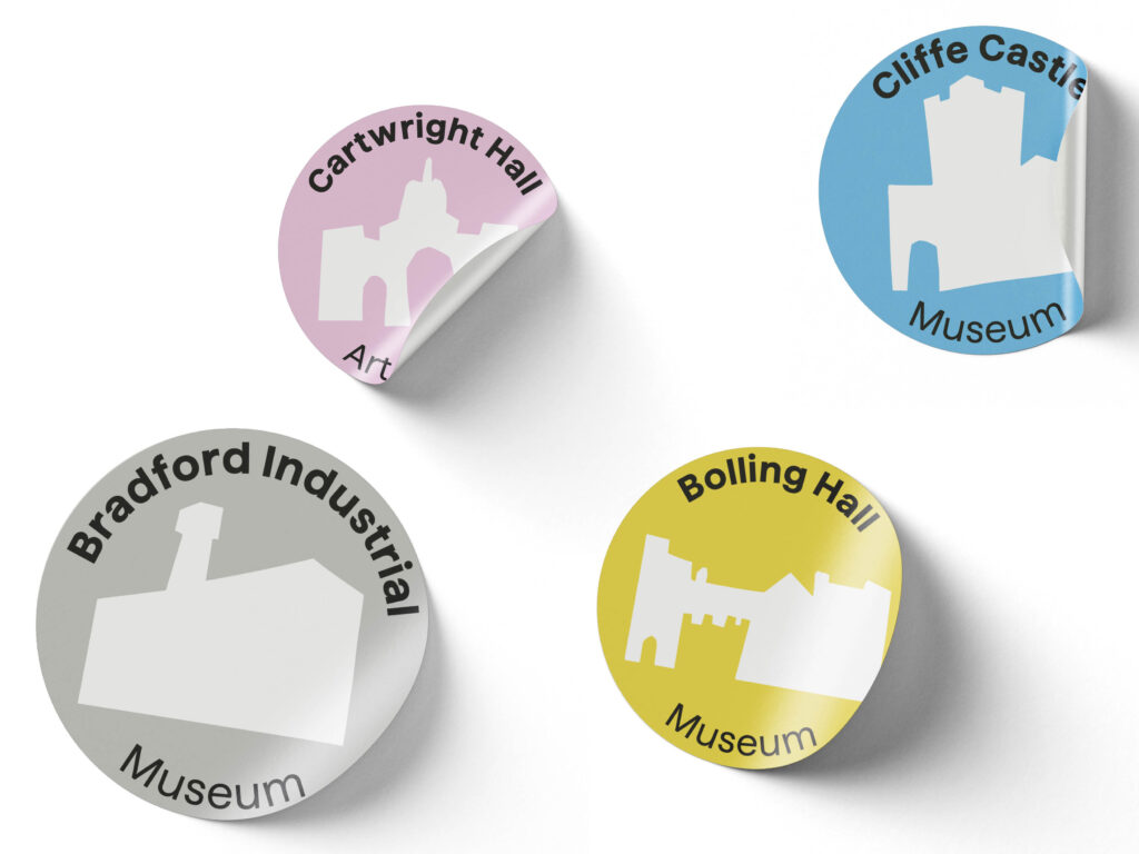 op left: Pink sticker with white silhouette of Cartwright Hall and text “Cartwright Hall Art”.
Top right: Blue sticker with white silhouette of Cliffe Castle and text “Cliffe Castle Museum”.
Bottom left: Grey sticker with white silhouette of Bradford Industrial Museum and text “Bradford Industrial Museum”.
Bottom right: Yellow sticker with white silhouette of Bolling Hall and text “Bolling Hall Museum”.
Each sticker has a curled edge effect, giving a 3D appearance.