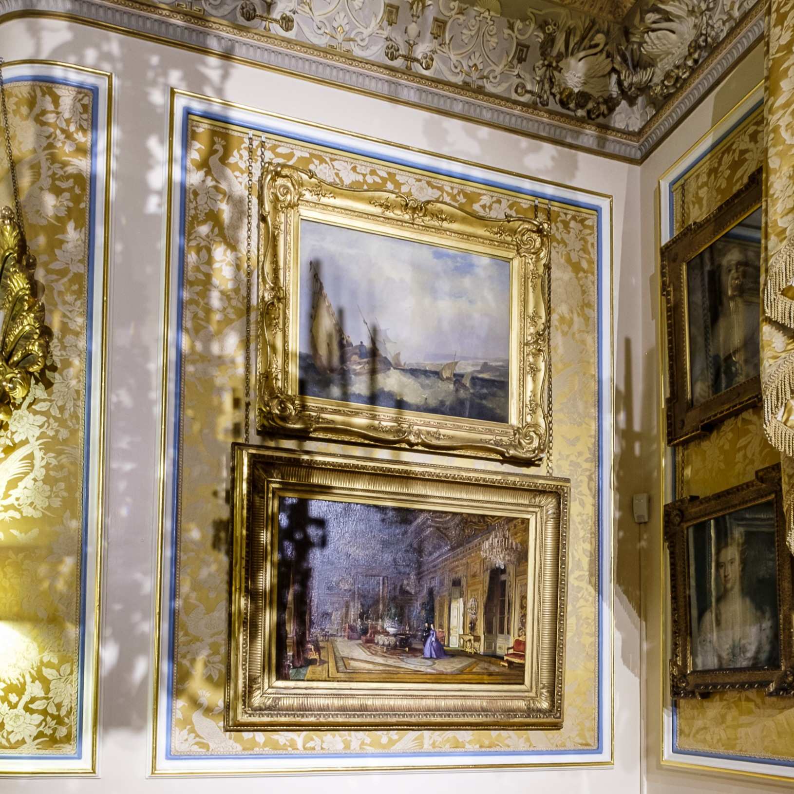 Interior wall of an ornate historic room with two large framed paintings in gilded frames. The upper painting depicts a misty landscape with trees and hills, while the lower painting shows an opulent interior scene with figures in period dress. The wall is decorated with gold floral patterns and blue trim, and the ceiling features elaborate plasterwork with leaf motifs. Shadows from decorative elements fall across the wall.
