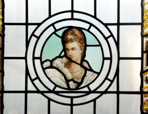 A stained glass window featuring a circular panel with a painted depiction of a person wearing an ornate white fur-trimmed garment and layered pearl necklaces. The figure has light brown hair styled neatly and adorned with a decorative gold headpiece. The background within the circle is a pale blue, and the surrounding glass is divided into geometric sections with black lead lines. Decorative yellow and gold details are visible along the edges of the window