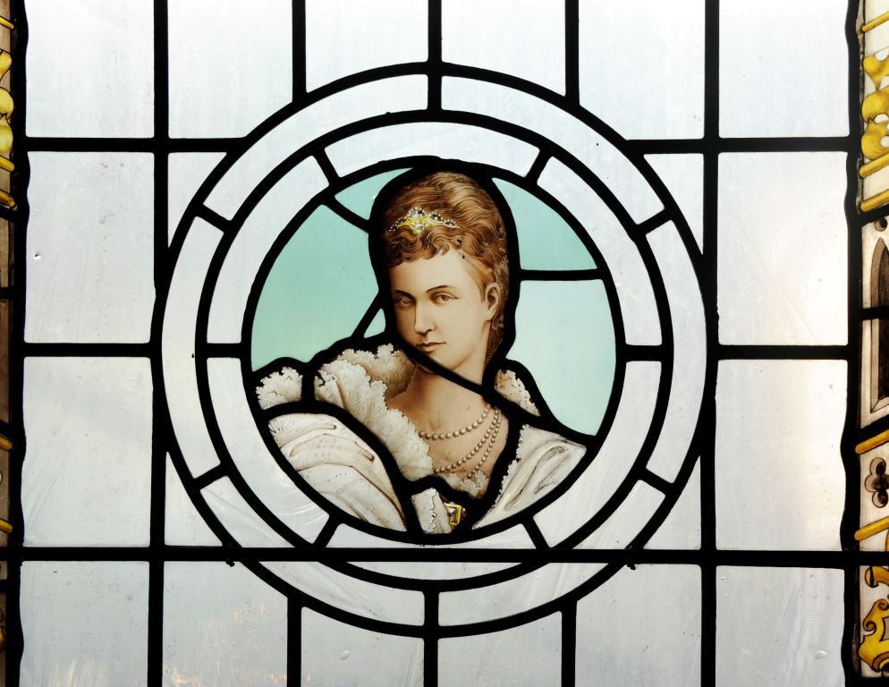A stained glass window featuring a circular panel with a painted depiction of a person wearing an ornate white fur-trimmed garment and layered pearl necklaces. The figure has light brown hair styled neatly and adorned with a decorative gold headpiece. The background within the circle is a pale blue, and the surrounding glass is divided into geometric sections with black lead lines. Decorative yellow and gold details are visible along the edges of the window