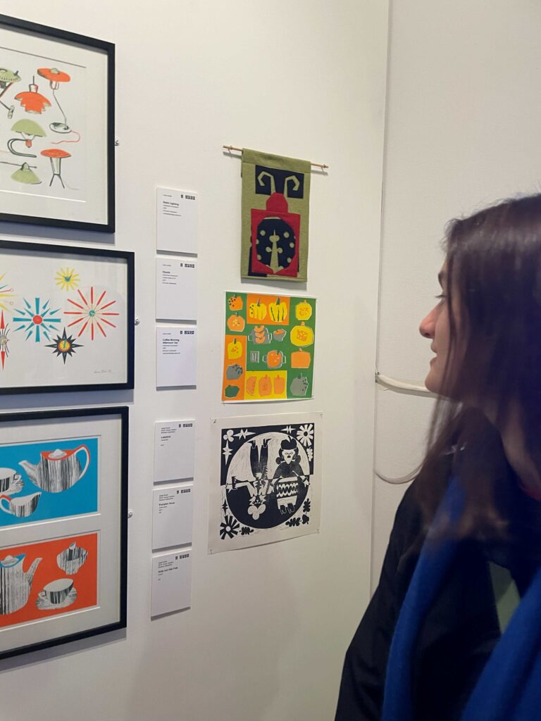 A close-up view of an art display on a white wall featuring several framed prints and textile pieces. On the left, three framed prints show colorful designs: one with abstract shapes, one with starburst patterns, and one with teapots and cups in bold colors. To the right, three smaller artworks are arranged vertically: a green textile piece with a stylized figure, a bright orange and green print with pumpkin motifs, and a black-and-white print with intricate patterns and figures. Exhibition labels with text and prices are visible next to the artworks.